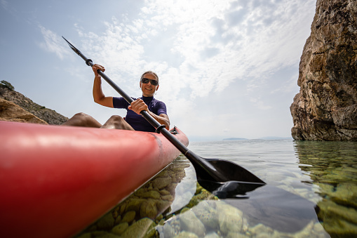 Fit athletic mature woman paddling in kayak during vacations in croatia smiling at camera, clear water with stones, cliffy coast on cloudy day