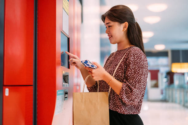 istock Smiling Asian woman holding smartphone and make mobile payment by using automated self service post terminal machine at street 1403615216