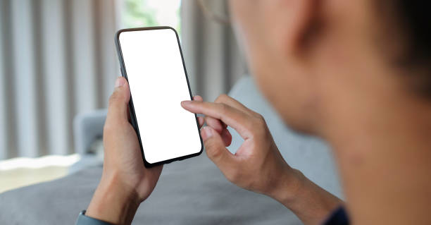 istock Young man hand holding smart phone while sitting on sofa at home. Over shoulder close up view, Blank screen for advertise. 1403618670
