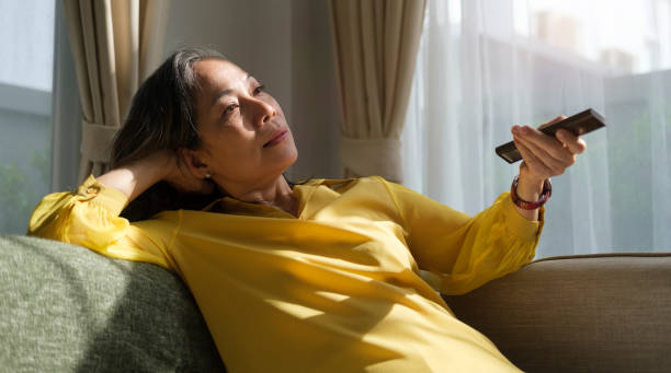 relaxed middle aged woman watching tv at home, sitting on couch with remote control. - changing channels imagens e fotografias de stock