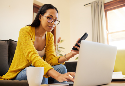Woman working remote while typing on her laptop and holding her smartphone sitting on a sofa in a bright living room. One focused young hispanic female with glasses at home using modern technology