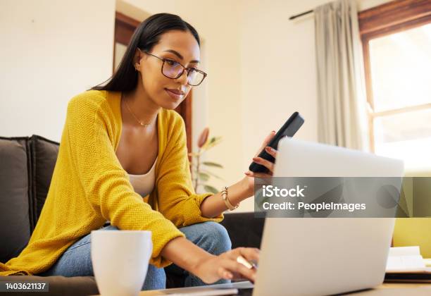 Woman Working Remote While Typing On Her Laptop And Holding Her Smartphone Sitting On A Sofa In A Bright Living Room One Focused Young Hispanic Female With Glasses At Home Using Modern Technology Stock Photo - Download Image Now