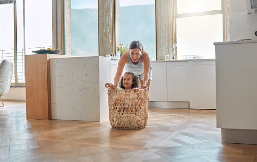 Young hispanic mother pushing her daughter around in a laundry basket at home. Young woman and her child playing and having fun while spending time at home