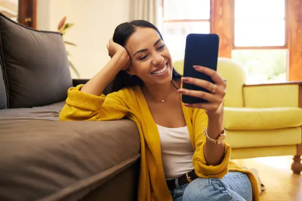 Photo of Woman smiling at her cellphone at home sitting on the floor against a sofa in a bright living room. A happy laughing young hispanic female video chatting on her smartphone on a call at home