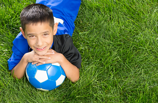 Smiling Mixed raced  8 years old boy wearing blue soccer uniform laying at soccer grass field.