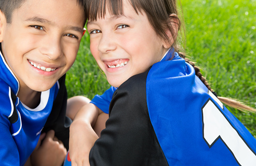 Smiling Mixed raced young sibling wearing blue soccer uniform sitting on soccer grass field.