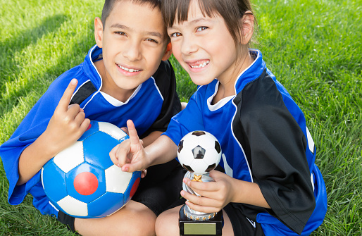 Smiling Mixed raced young sibling celebrating winning wearing blue soccer uniform sitting on grass.