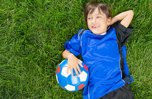 Above view of Smiling Mixed raced  8 years old girl wearing blue soccer uniform laying on grass soccer field looking away.