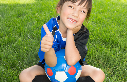 Smiling Mixed raced  8 years old girl wearing blue soccer uniform sitting on soccer grass field gesturing thumbs up.