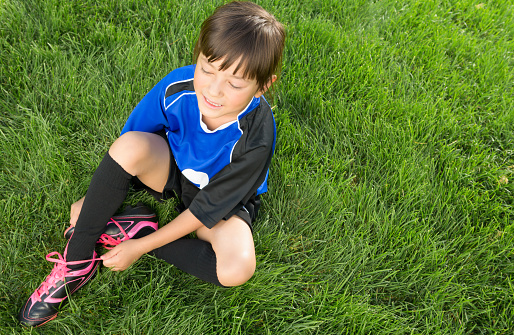 Smiling Mixed raced  8 years old girl wearing blue soccer uniform sitting on soccer grass field fixing her shoelace.