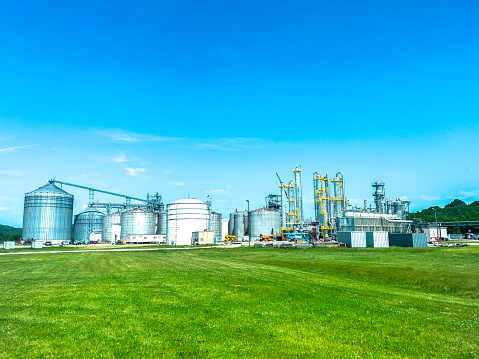 An Ethanol refinery plant is surrounded by a green field and blue sky. The image features silos and buildings surrounded by lush greenery.