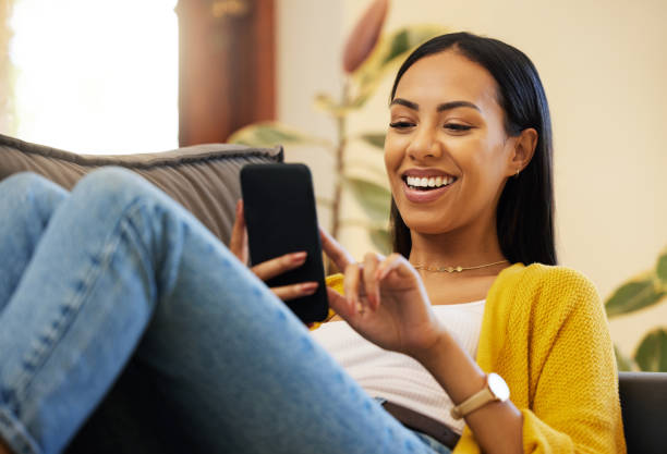 istock Hispanic woman using her smartphone while smiling and relaxing in a bright living room. A young female lying on a sofa scrolling on her cellphone at home using modern technology during lockdown 1403641079