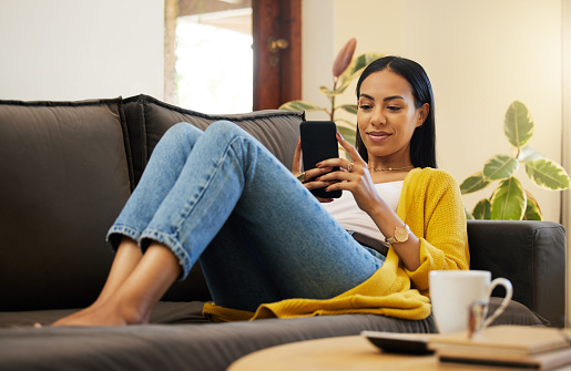 Hispanic woman using her smartphone while comfortable and relaxing in a bright living room. A young female lying on a sofa texting on her cellphone at home using modern technology during lockdown
