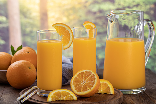 Two glasses and pitcher of orange juice with sliced oranges against a rural summer background