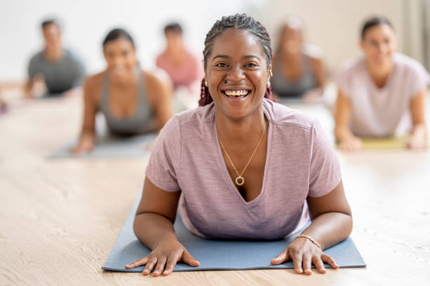 A young woman of African decent lays out on her stomach on a yoga mat as she holds a cobra pose.  She is dressed comfortably in athletic wear and surrounded by her peers as they participate in the class together.