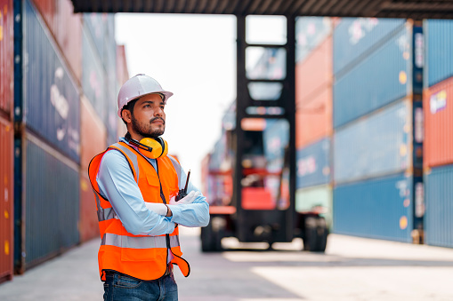 A dock worker with a clipboard and pen at a port with a cargo ship and crane in the background.