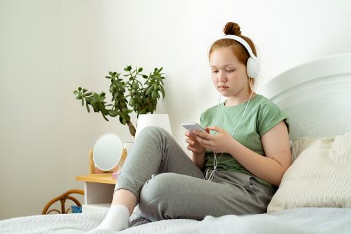 Caucasian teenage girl with headphones is sitting on bed with smartphone, smiling, watching videos on Internet, listening to music, communicating with friends on social networks