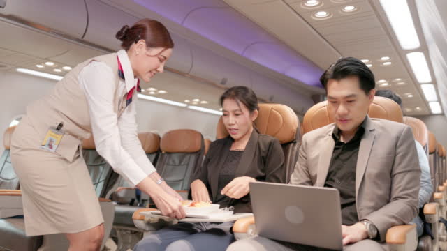 On the plane, a friendly Asian female flight attendant serves food and drinks while conversing with customers. Airline service is available.