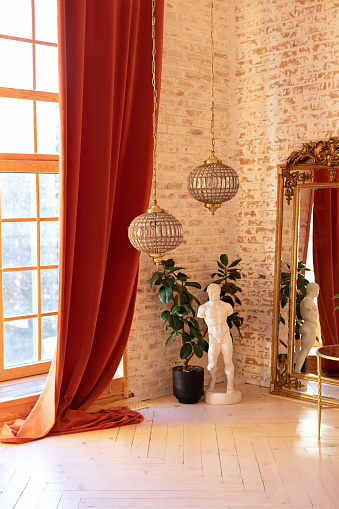 Modern living room in French style. Stylish French interior with big window and curtains, green plant in pot, classical sculpture, vintage gold mirror and crystal chandeliers against a brick wall.