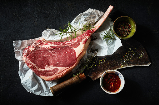 Top view of uncooked tomahawk steak with spices placed near chopping axe on baking parchment on black background