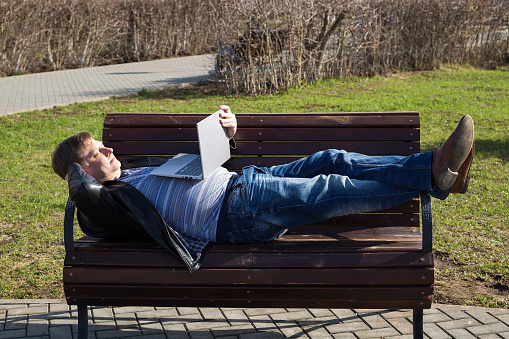 An adult man lay down on a bench in the park and watches a movie on a laptop.