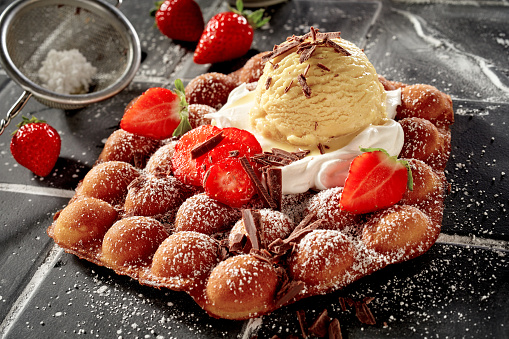 Gourmet bubble waffle summer dessert with fresh strawberries, whipped cream and fruity berry ice-cream topped with flaked chocolate and icing sugar served on tiles for menu advertising