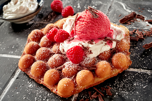 Delicious bubble waffle topped with raspberry ice cream and fresh ripe red berries garnished with chocolate flakes on a bed of whipped cream