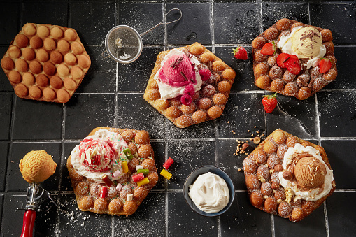 Top down on an assortment of bubble waffles with fresh fruit and nutty ice-cream on a bed of whipped cream drizzled with chocolate shavings and icing sugar on grey tiles