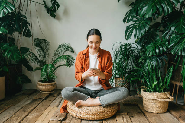 a beautiful young woman is sitting in a lotus position in a beautiful green garden among plants, turning off her phone or choosing music for meditation. the concept of mindfulness, psychological and mental health. - prayer call imagens e fotografias de stock