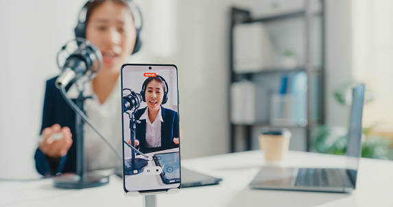 Close up of young Asian businesswoman recording content with smartphone and broadcasting a podcast on her laptop from studio office. Live streaming and broadcasting online concept.