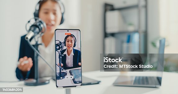 istock Close up of young Asian businesswoman recording content with smartphone and broadcasting a podcast on her laptop from studio office. 1403684204