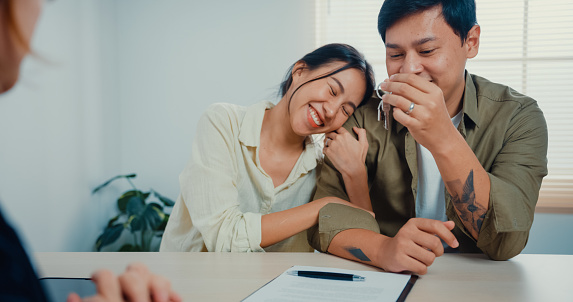 Saleswoman keep house key to young married couple with casual sitting at desk feeling happy with new home contract agreement at estate office.