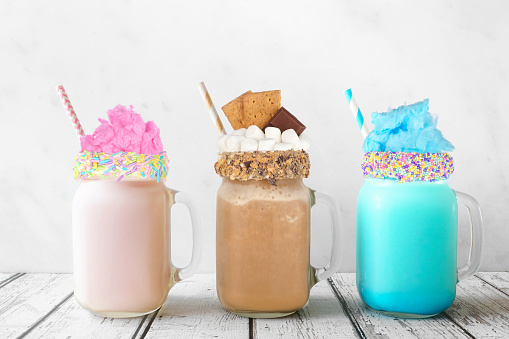 Fun colorful summer milkshakes in mason jar glasses against a white background. Blue and pink cotton candy and chocolate smores sweet drinks.