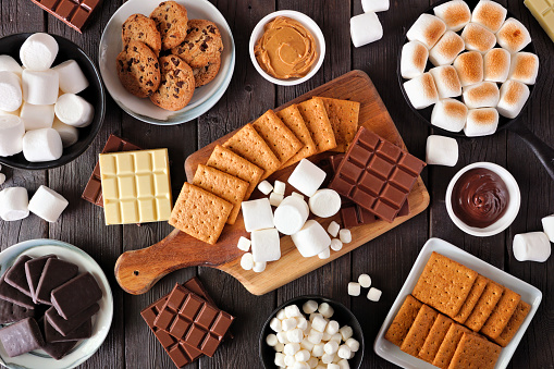 Smores buffet table scene. Roasted marshmallows, crackers, chocolate and an assortment of ingredients. Top down view over a dark wood background.