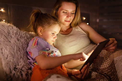 Night time mother and her young daughter laying in bed, using a digital tablet