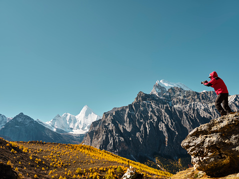 asian man standing on top of rock taking photo of Yangmaiyong (or Jampayang in Tibetan) mountain peak in the distance in Yading, Daocheng County, Sichuan Province, China