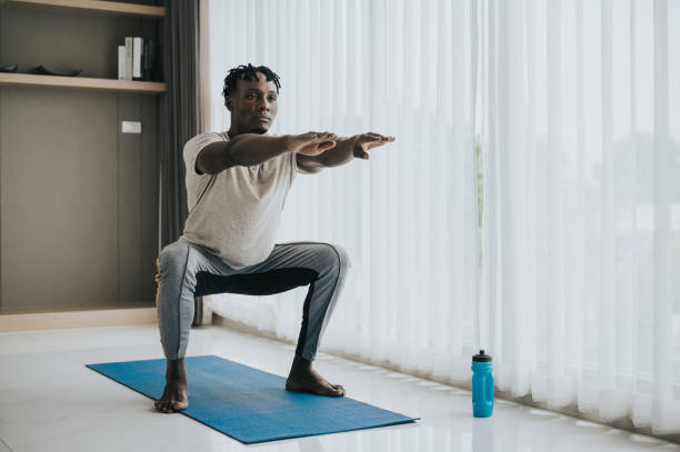 African man doing squat exercise African American man doing squat exercise during workout in livingroom on yoga mat at home crouching stock pictures, royalty-free photos & images