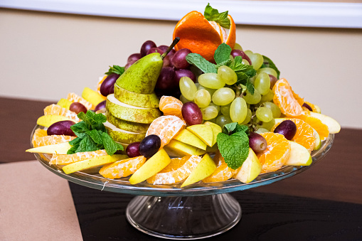 Fruit plate. Stylishly lined fruit: orange slices, pear slices, grapes, tangerine, mint leaves. Catering serving on a glass base.