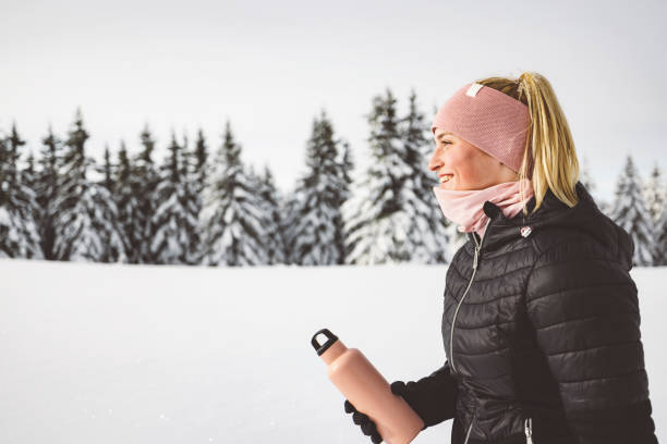 Side profile of a smiling blonde woman with her hair in a ponytail holding a pink thermo flask in her hands. Winter forest in the background. Winter hike.