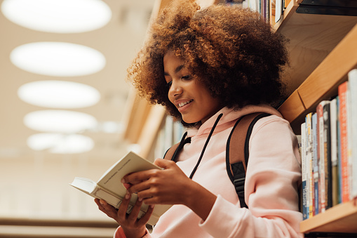 Chica en casuales con mochila inclinada estantería en biblioteca. Estudiante leyendo un libro mientras está de pie en la biblioteca. photo