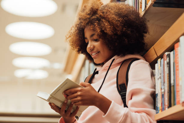 girl in casuals with backpack leaning bookshelf in library. student reading a book while standing in library. - lido imagens e fotografias de stock