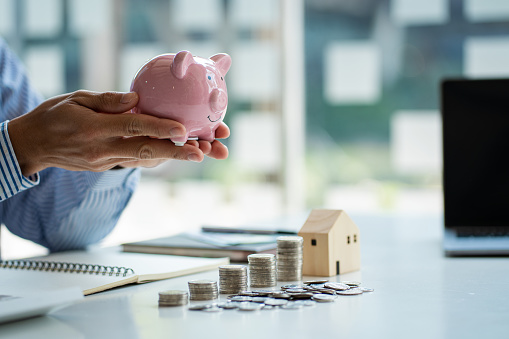 Businessman hand holding a piggy bank with piles of coins and a wooden house on the table, future house savings concept.