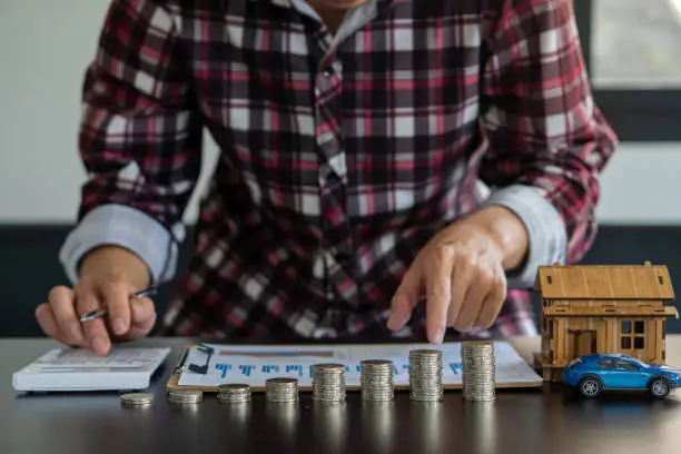 Photo of man hand holding pen press calculator pointing financial graph with pile of coins in front with car and small house on table financial accounting concept