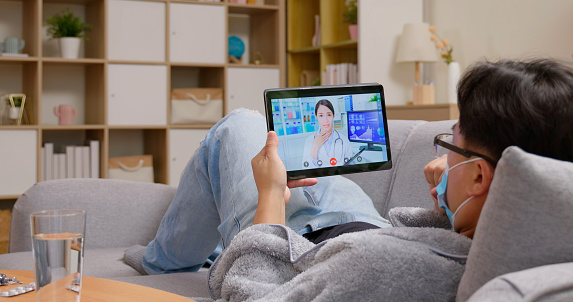 asian male sick patient wear mask lying on sofa use tablet has telemedicine video call with female doctor at home
