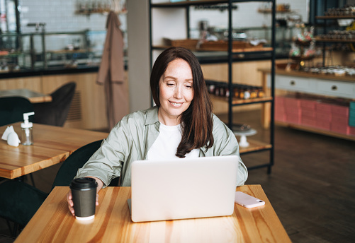 Adult smiling brunette business woman forty years with long hair in stylish shirt working on laptop using mobile phone in cafe