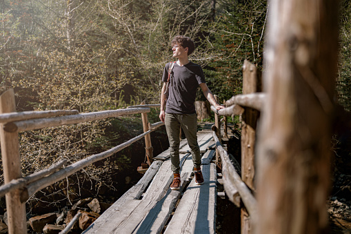 Young handsome guy standing on old wooden bridge in mountains forest. Summer tourism. Traveling.