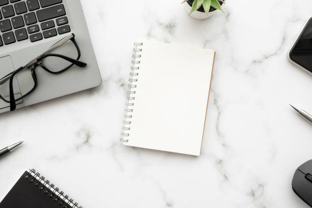 istock Blank notebook is on top of white marble office desk table with laptop computer and office supplies. Top view with copy space, flat lay. 1403788478