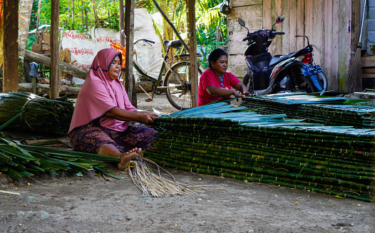 Blang Pidie, Indonesia - January 24, 2022: Marinah while weaving Rumbia leaves that will be made roofing instead of tiles. Until now, this rumbia roof leaf is still in great demand by business actors.  although it currently competes with modern roofs. This rumbia roof leaf is used for traditional home needs, as well as tourist attraction camps. This rumbia roof leaf is sold at a price range of Rp.5000 - Rp.7.000