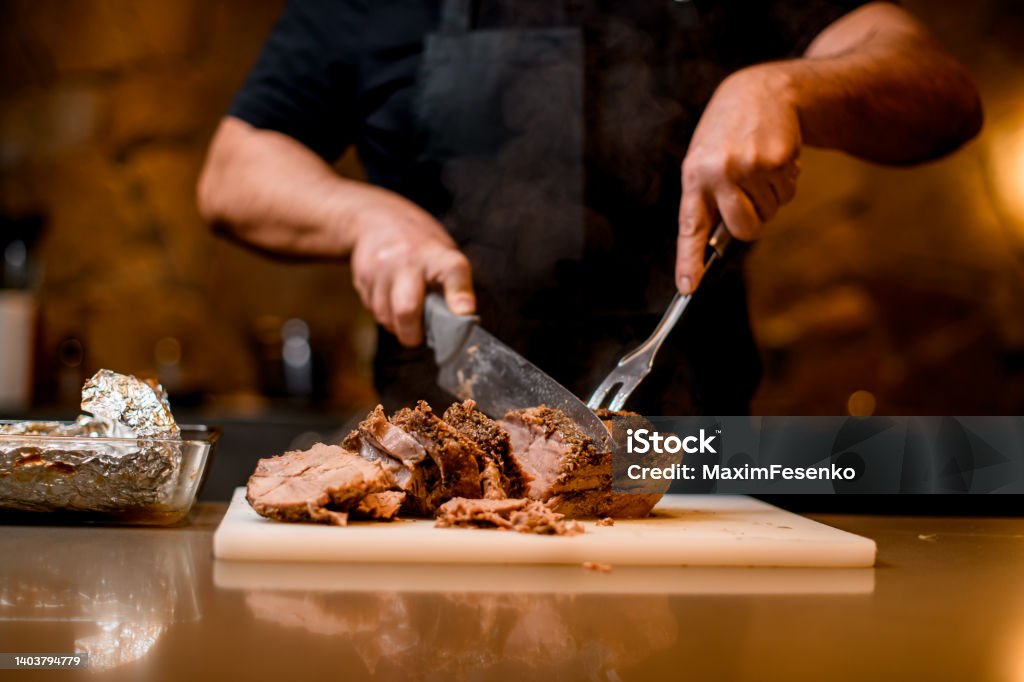 view on fried meat which man cuts to slices on cutting board. view on tasty hot fried meat which man chef masterfully cuts to slices on white cutting board. Barbecue - Meal Stock Photo