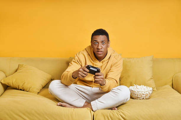 istock Concentrated man playing video games while sitting on the couch with yellow background 1403798831